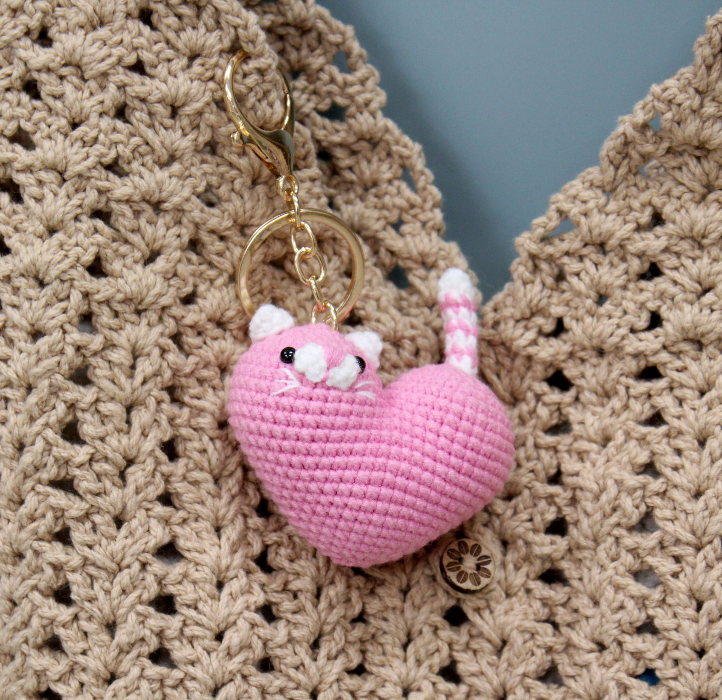 Pink and white crochet heart-shaped cat keychain with gold hardware, resting on a crocheted fabric.