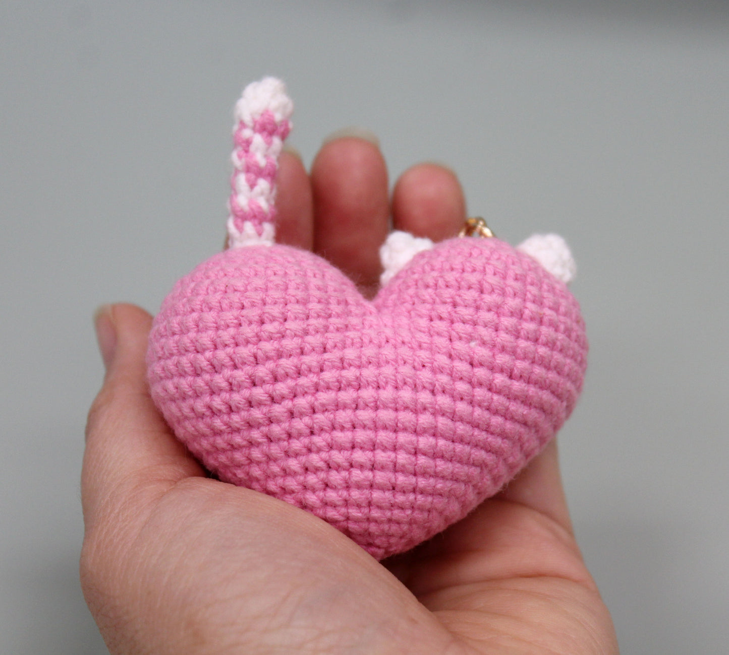 Pink and white crocheted heart-shaped keychain with cat ears and a tail, held in hand.