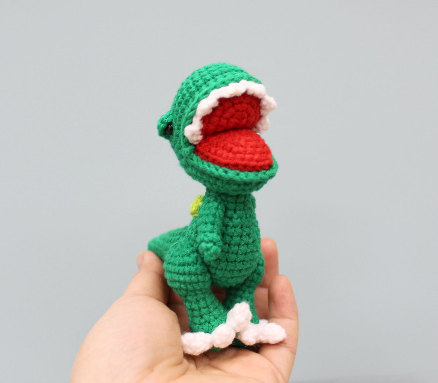 A green crocheted dinosaur keychain with a red mouth, white teeth, and pink feet.