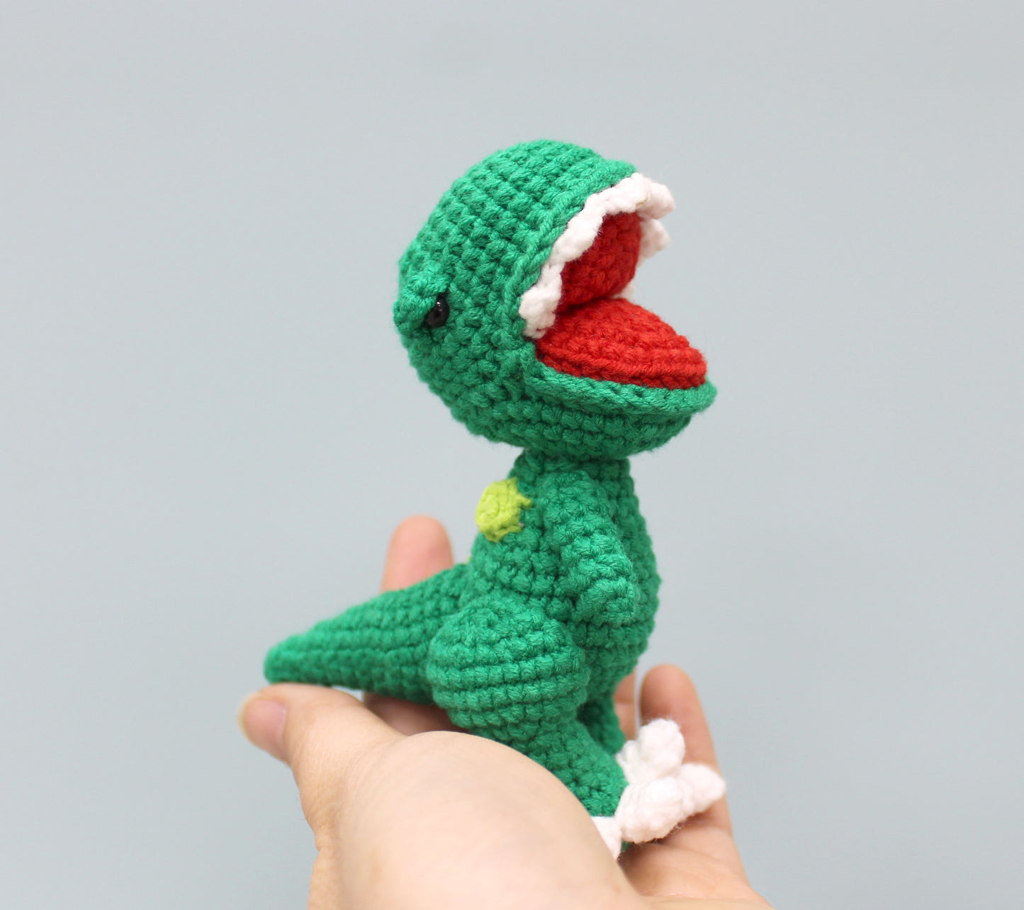 A green crocheted dinosaur keychain with a red mouth, white teeth, and a small yellow spot on its back.