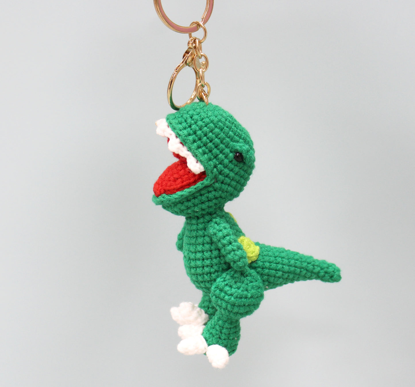 A green crocheted dinosaur keychain with a red mouth, white teeth, and a yellow spot on its back.