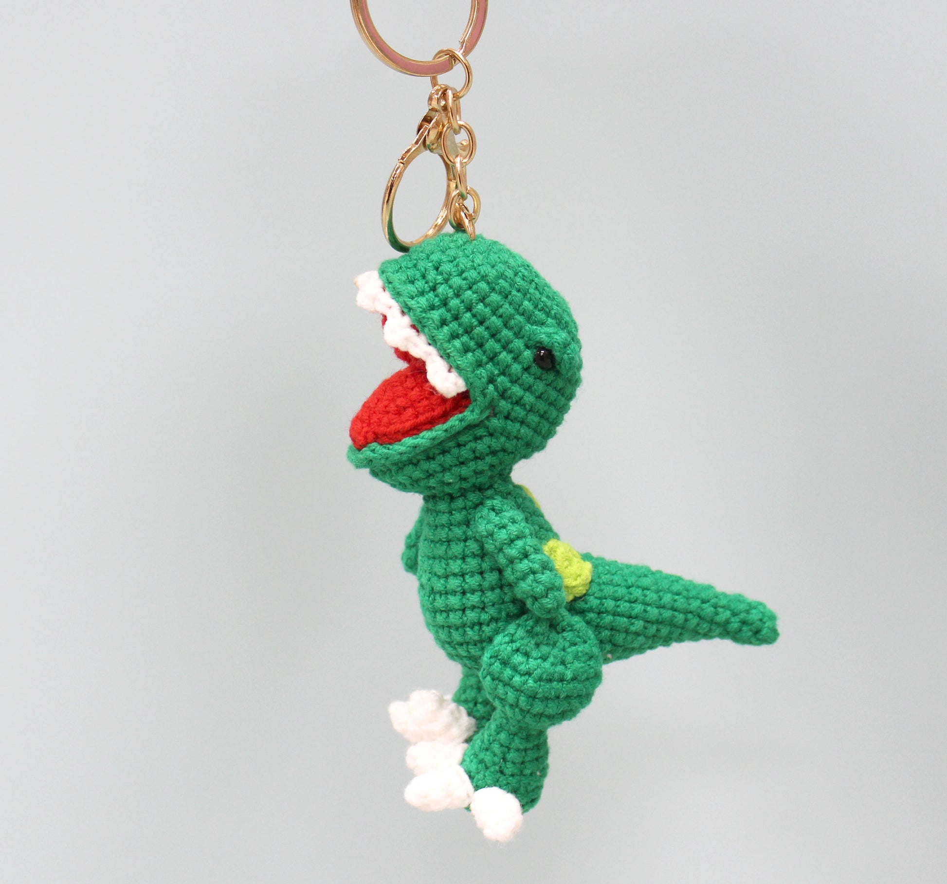 A green crocheted dinosaur keychain with a red mouth, white teeth, and a yellow spot on its back.