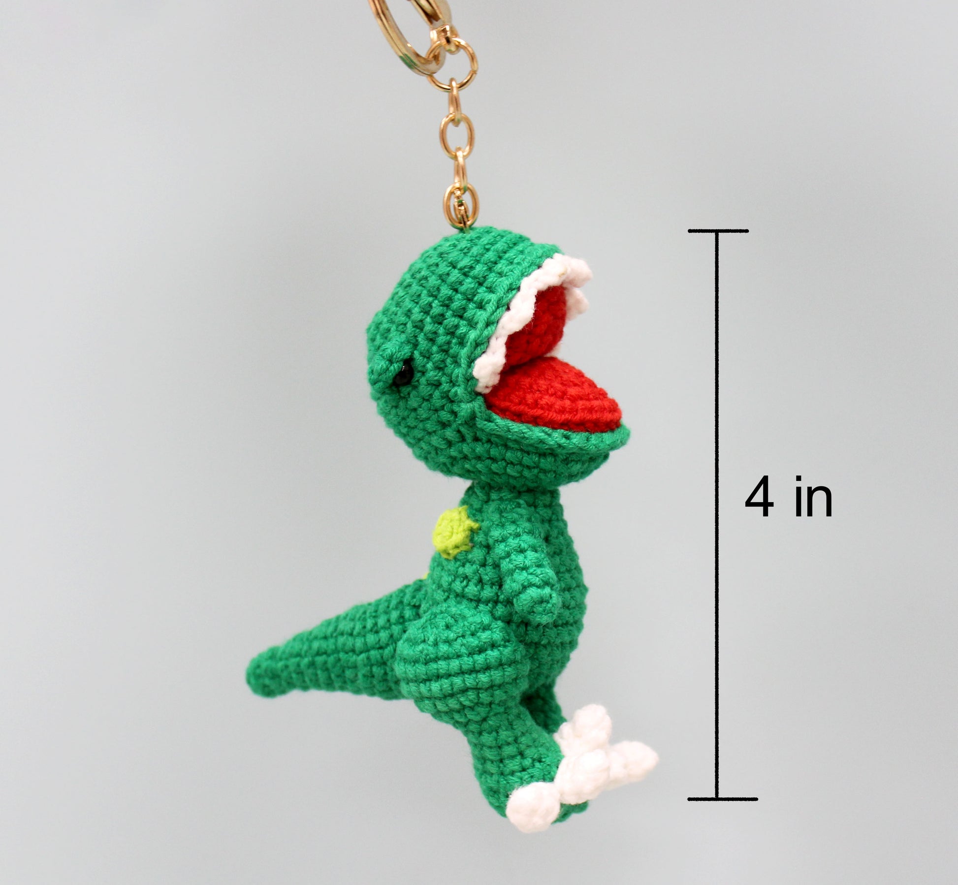 A green crocheted dinosaur keychain with a red mouth, white teeth, and a yellow spot, measuring 4 inches tall.