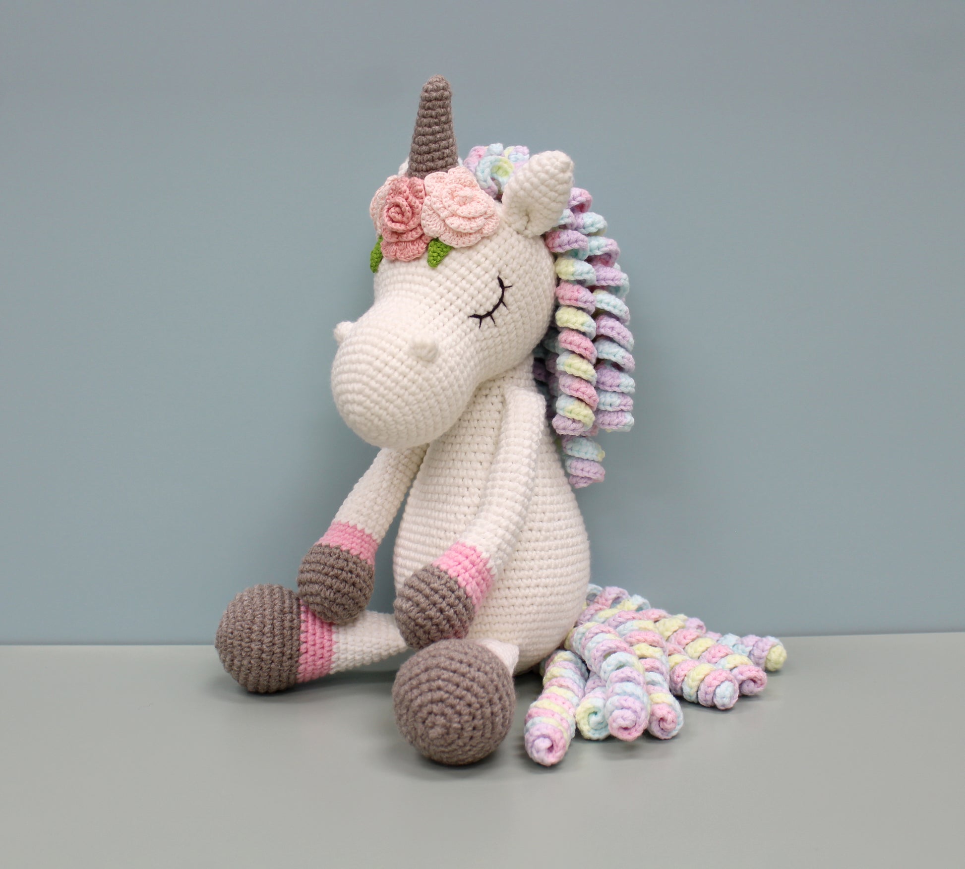 Handmade crochet unicorn plush with multicolored mane and floral headpiece, seated and smiling.