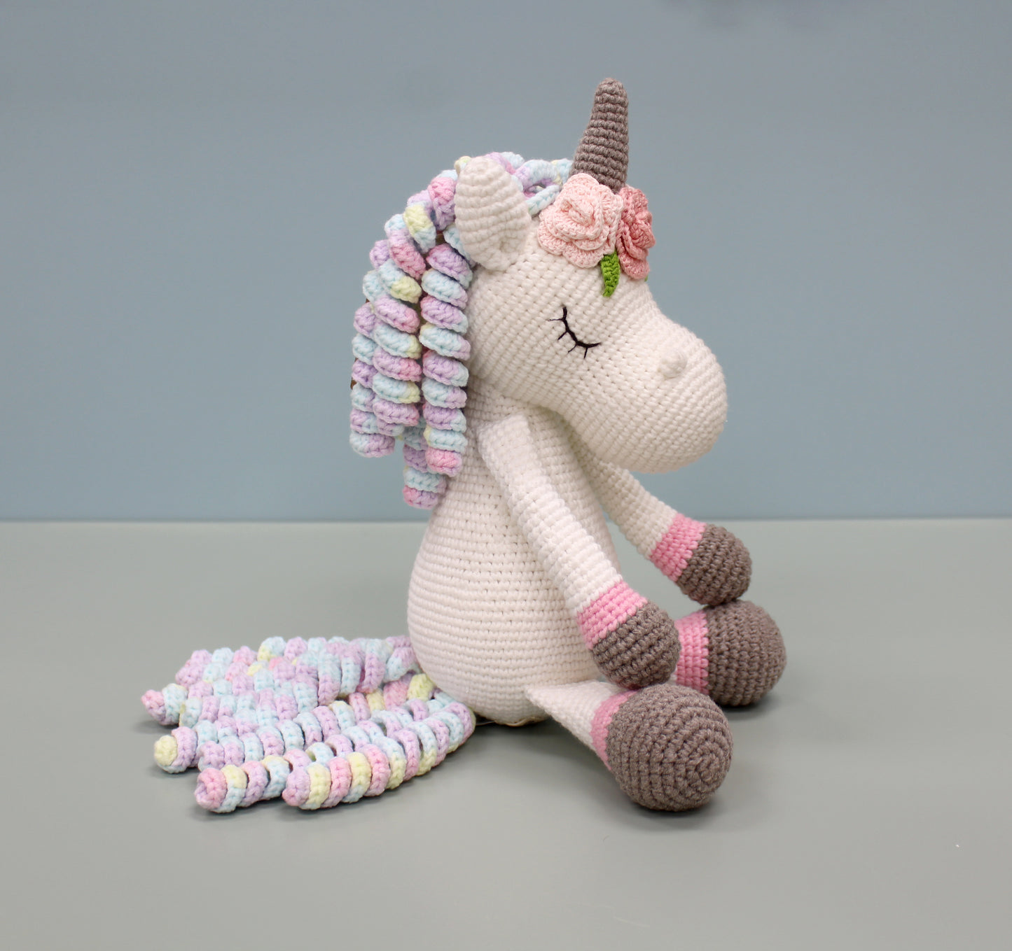 Handmade crochet unicorn plush with multicolored mane and tail, featuring a brown horn and floral ear accents.
