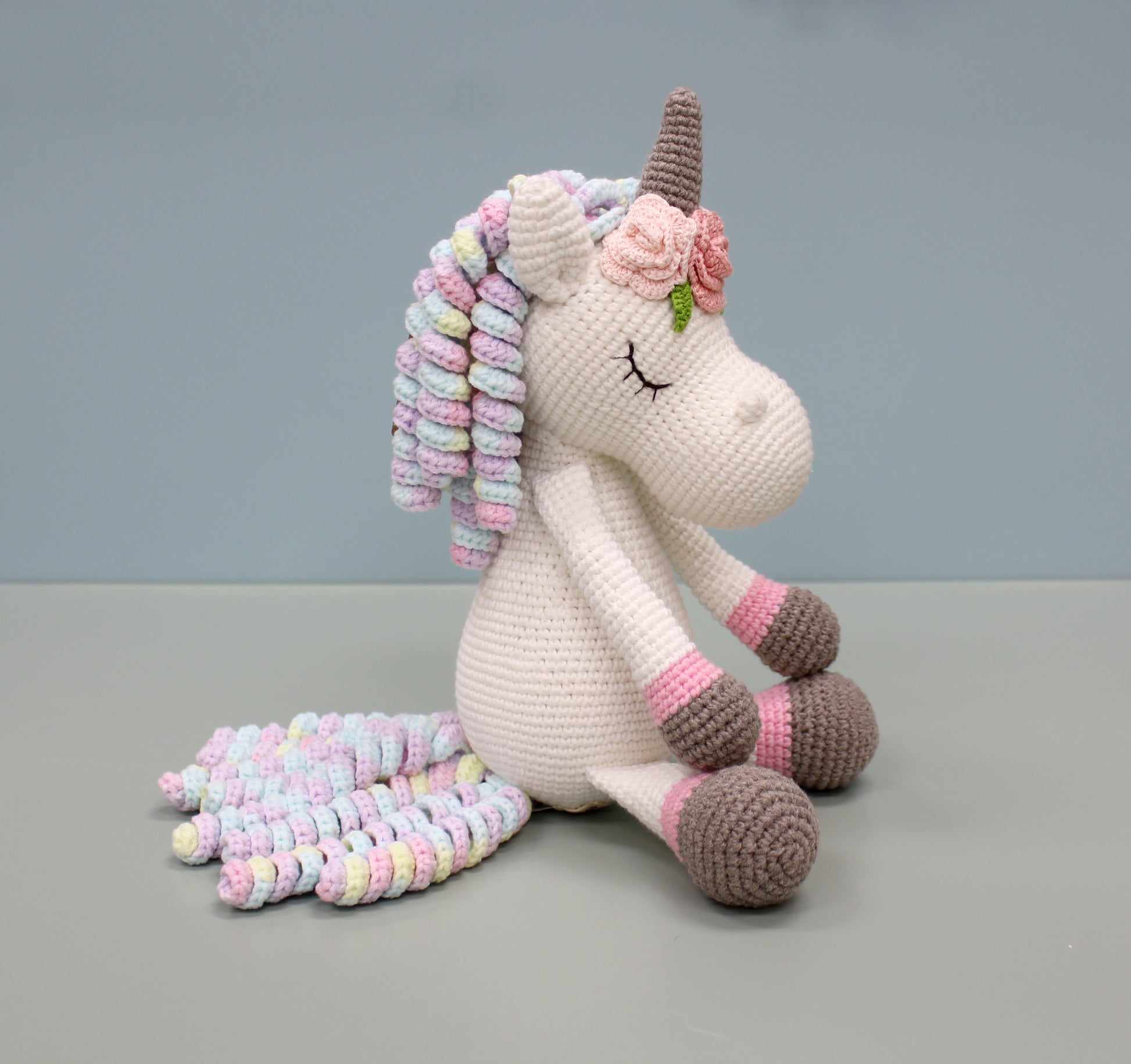 Handmade crochet unicorn plush with multicolored mane and tail, featuring a brown horn and floral ear accents.