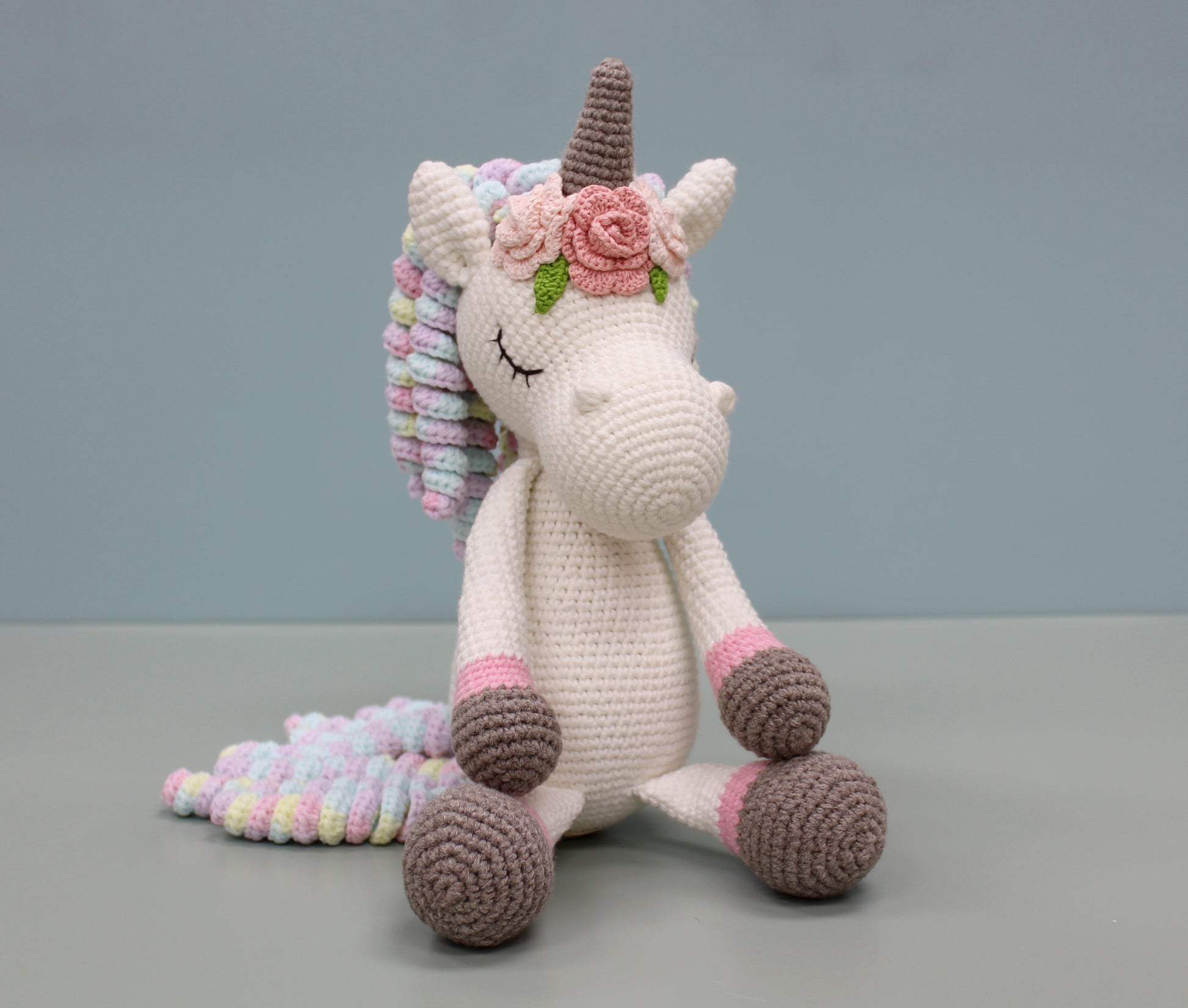 Handmade crochet unicorn plush with multicolored mane, pink floral adornment, and soft pastel accents.