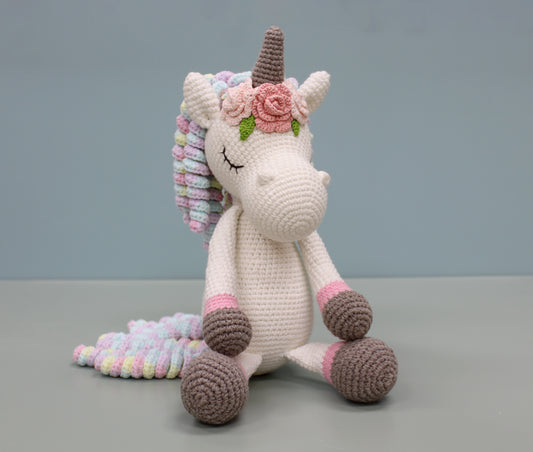 Handmade crochet unicorn plush with multicolored mane, pink floral adornment, and soft pastel accents.