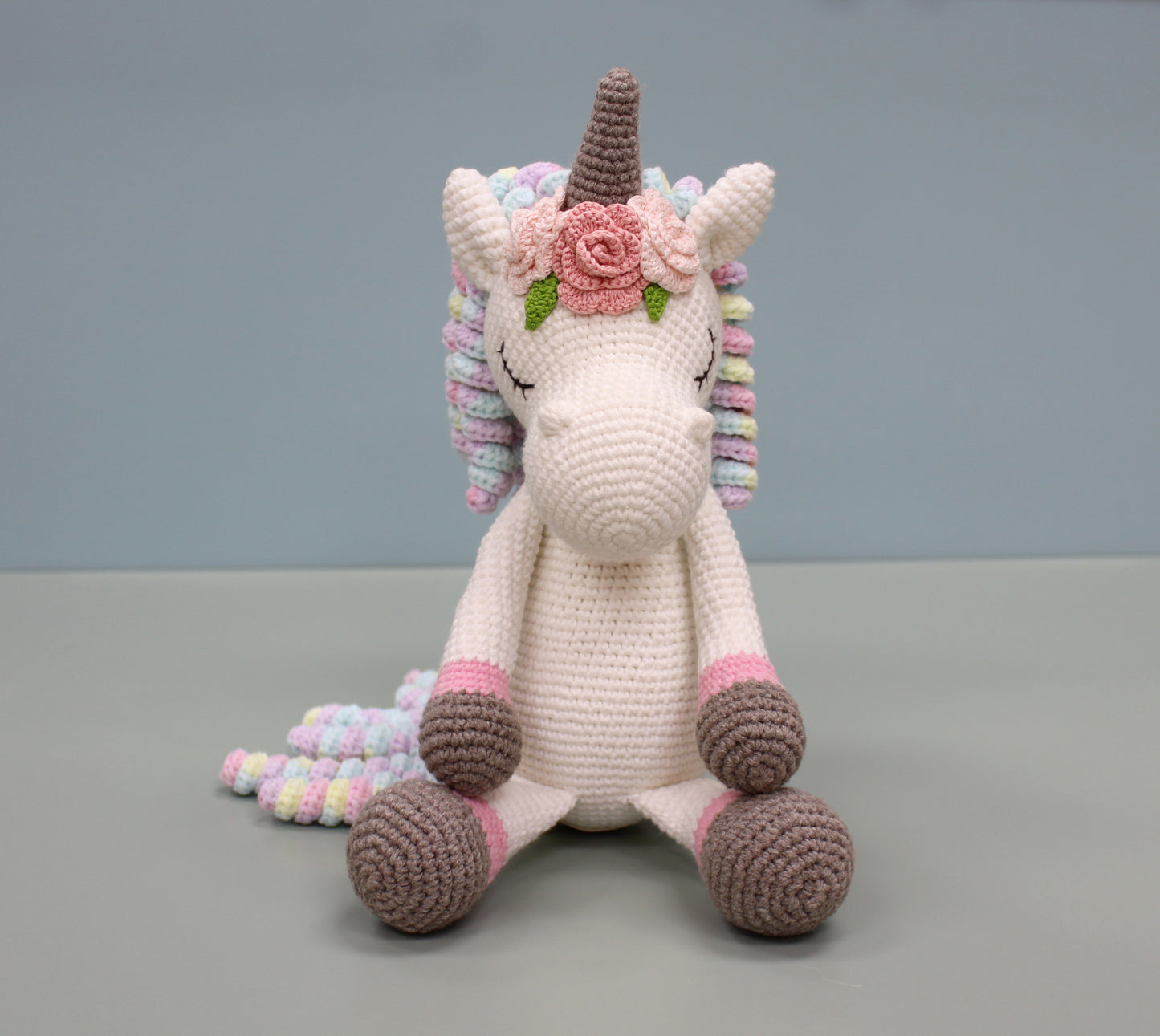 Handmade crochet unicorn plush with multicolored mane, pink floral adornment, and brown-and-pink hooves.