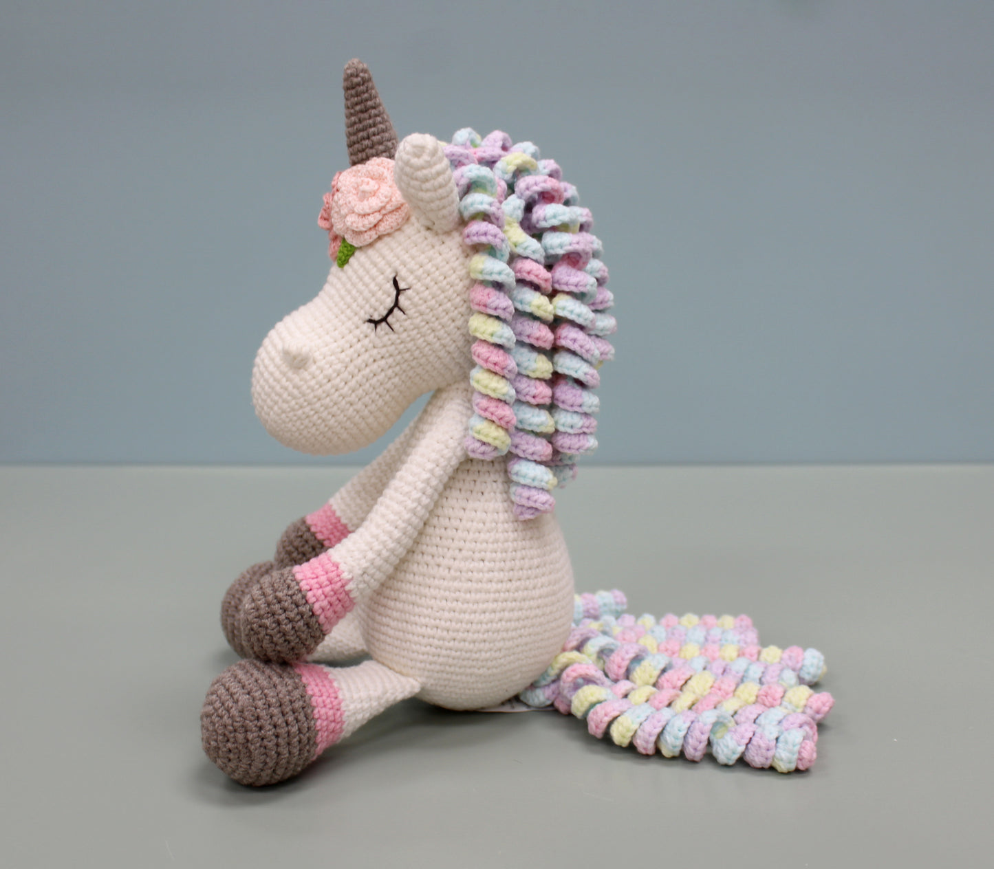 Handmade crochet unicorn plush with multicolored mane and tail, featuring a brown horn and gentle smile.