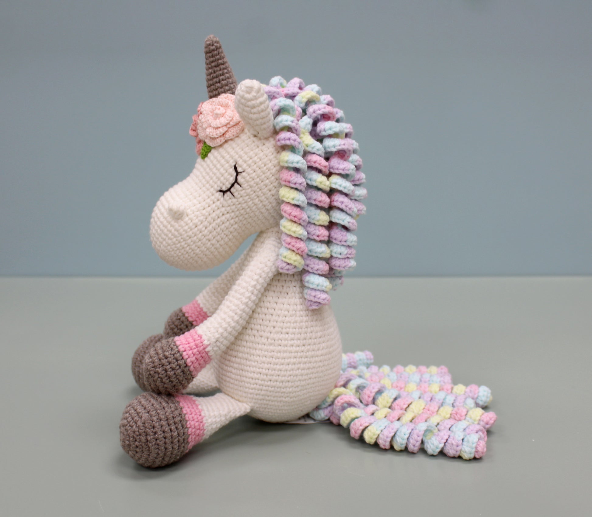 Handmade crochet unicorn plush with multicolored mane and tail, featuring a brown horn and gentle smile.