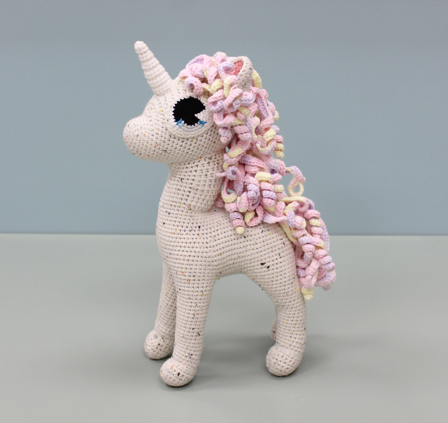 Handmade crochet unicorn stuffed animal with a colorful, curly pastel mane and a speckled cream body.
