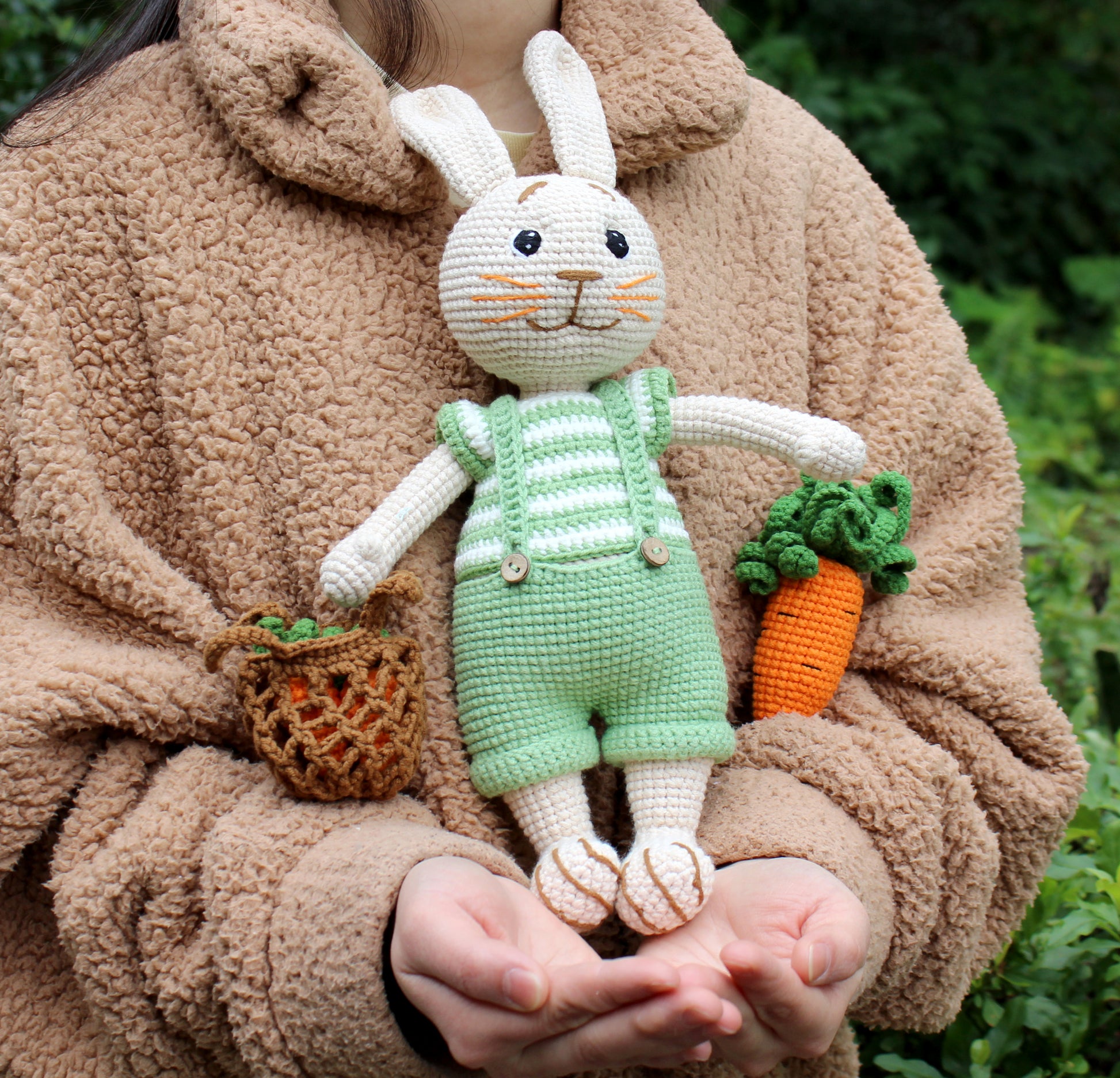 Handmade crochet Easter bunny in green and white overalls with a carrot and basket, held gently.
