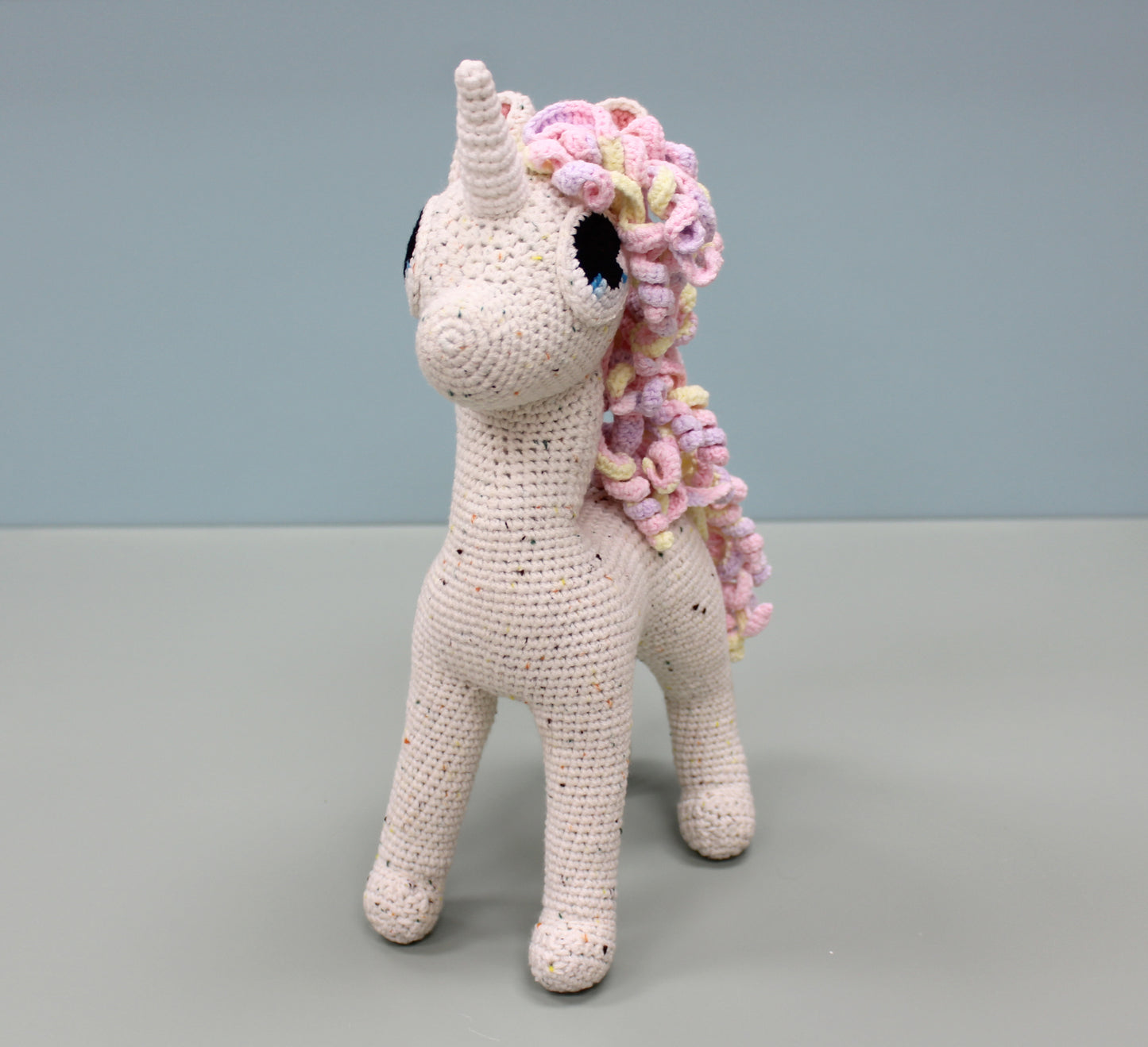 Handmade crochet unicorn stuffed animal with speckled white body, colorful pastel mane, and horn.