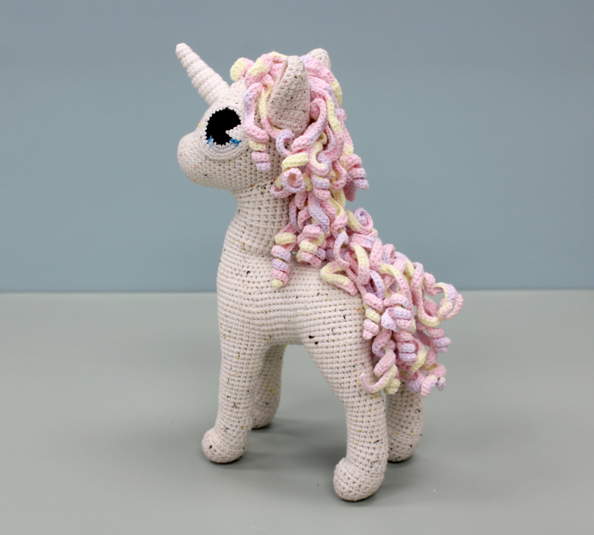Handmade crochet unicorn plush with multicolored speckled body and pastel pink and yellow spiral mane and tail.