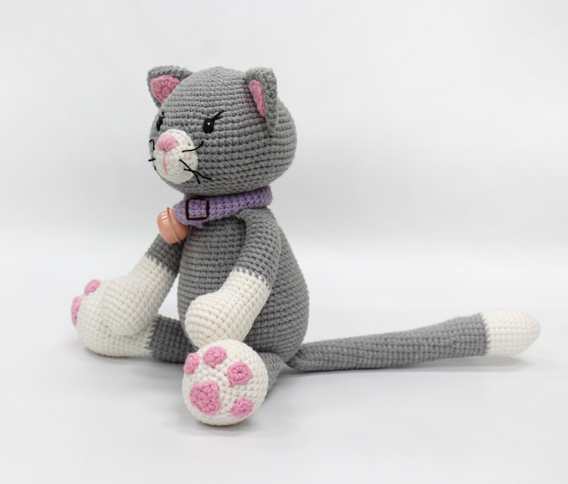 Gray and white crochet stuffed cat with pink paws and a purple collar, posed upright.