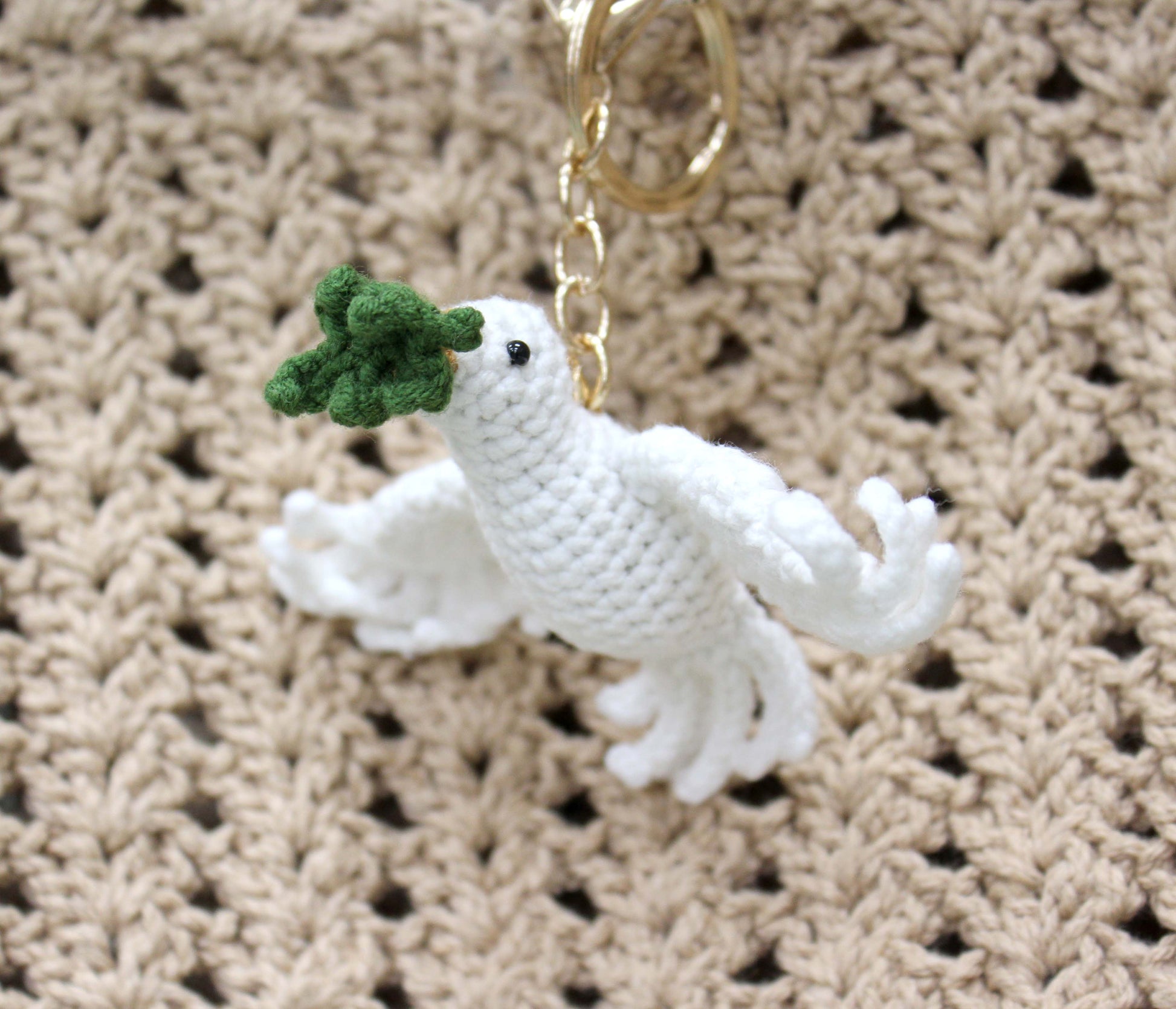 Handmade white crochet dove keychain holding a green floral detail, attached to a gold ring.