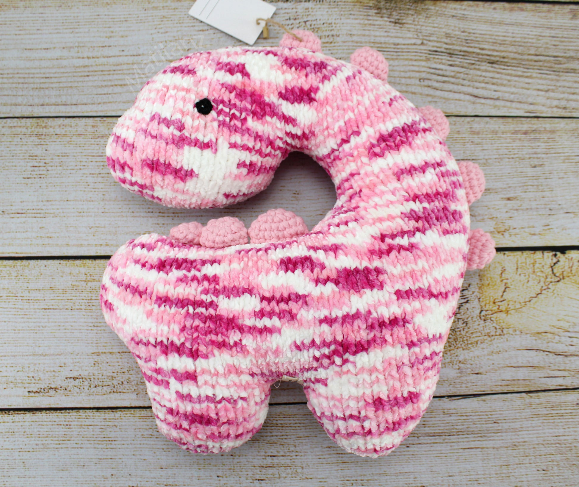 Handmade pink and white striped crochet dinosaur neck pillow with a U-shaped design and small spikes.