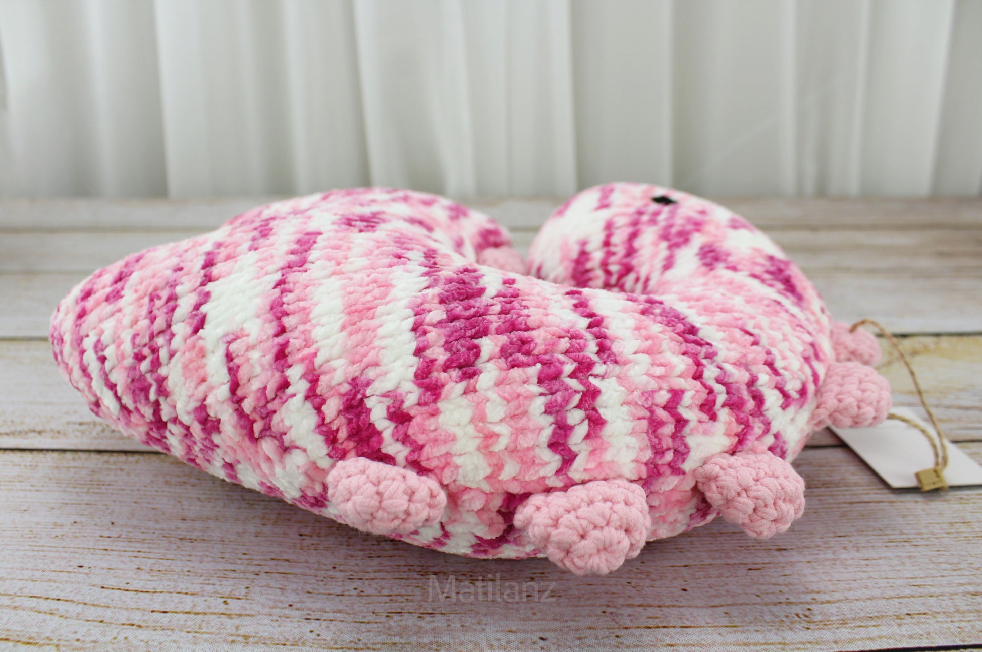 Handmade crochet dinosaur neck pillow in pink and white with pom-pom legs and a tag.