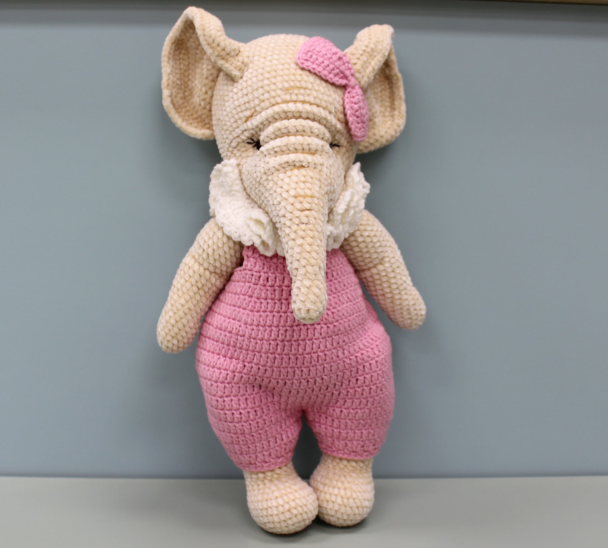 "Adorable crochet elephant toy wearing pink overalls and matching bow, standing upright."