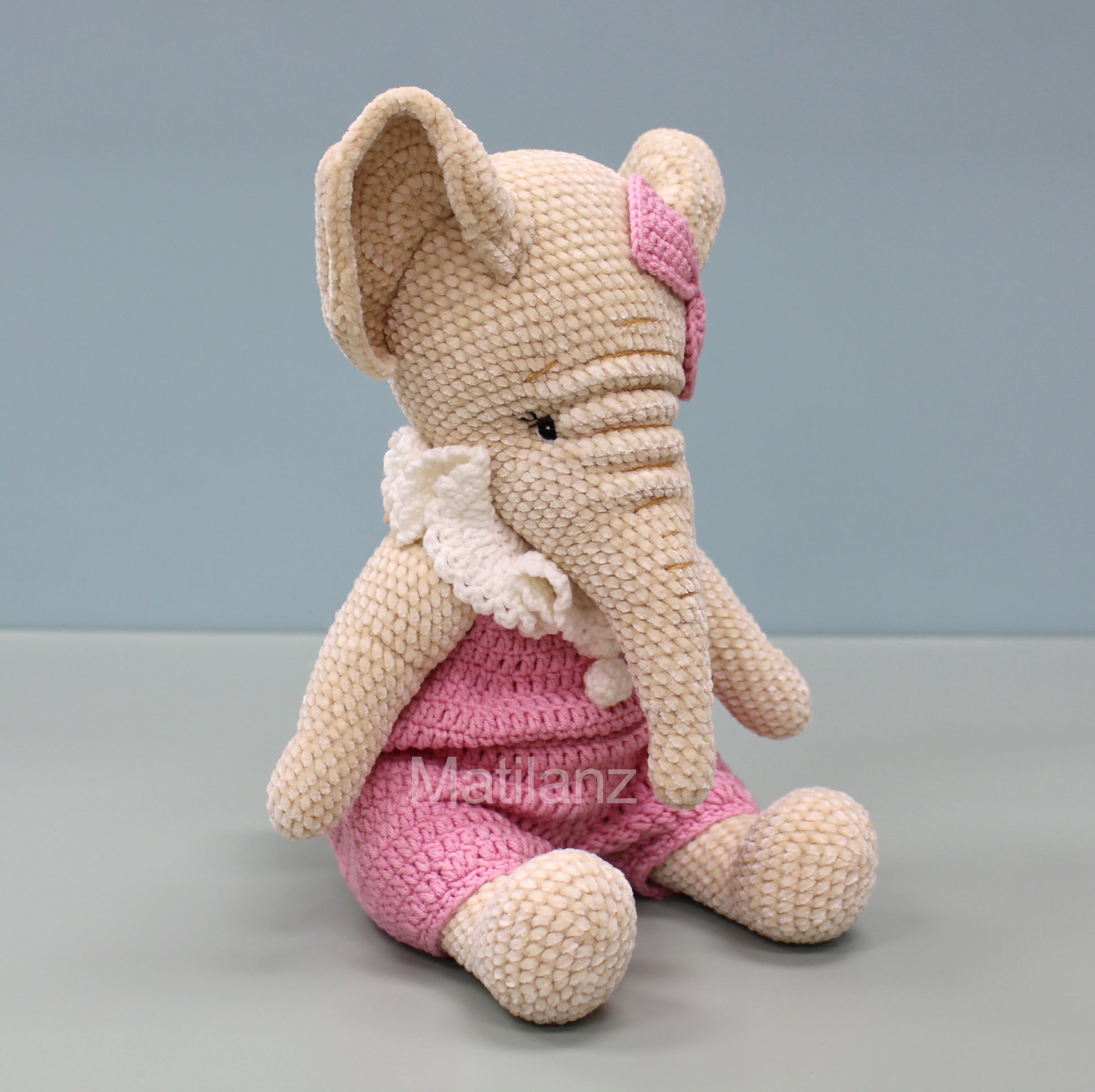 Crochet elephant in pink overalls, 13 inches tall, with a white bow and pink ear detail.