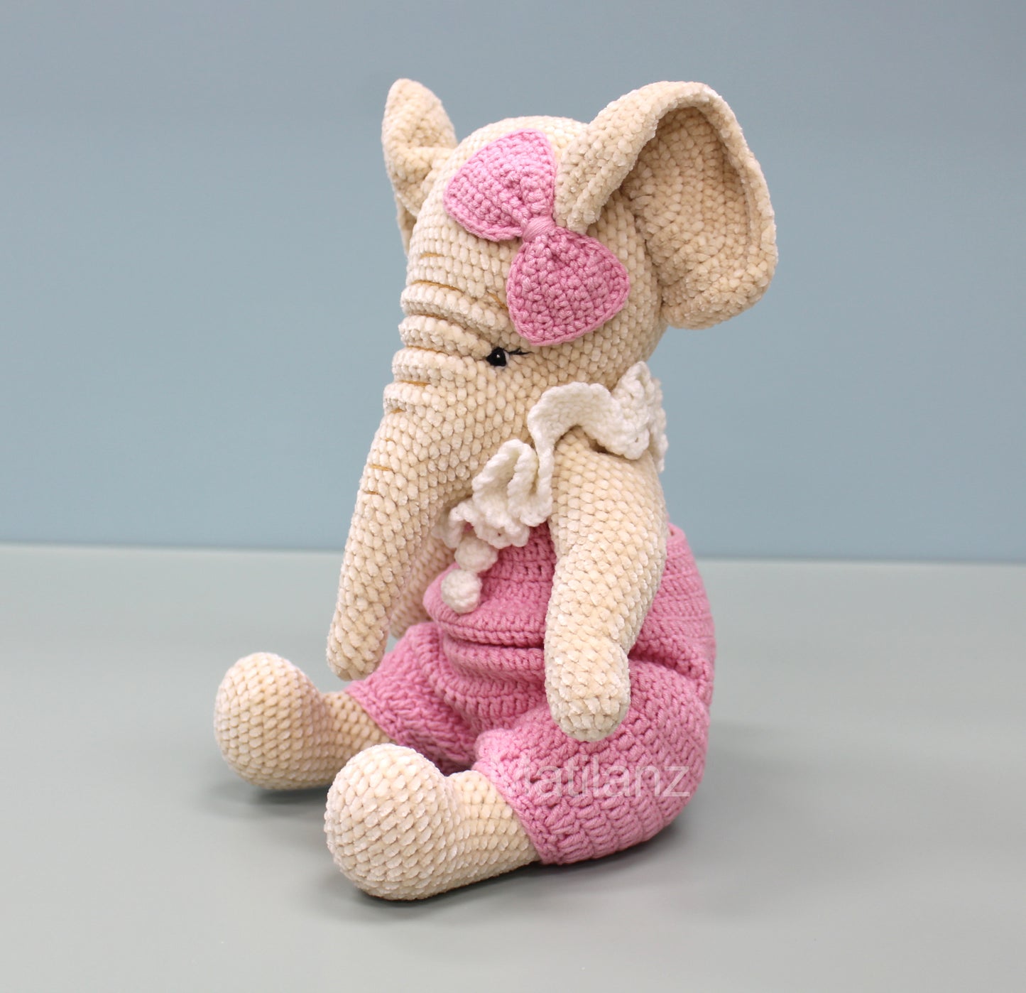 A handmade crochet elephant wearing a pink dress and bow, sitting with its trunk resting on its knee.