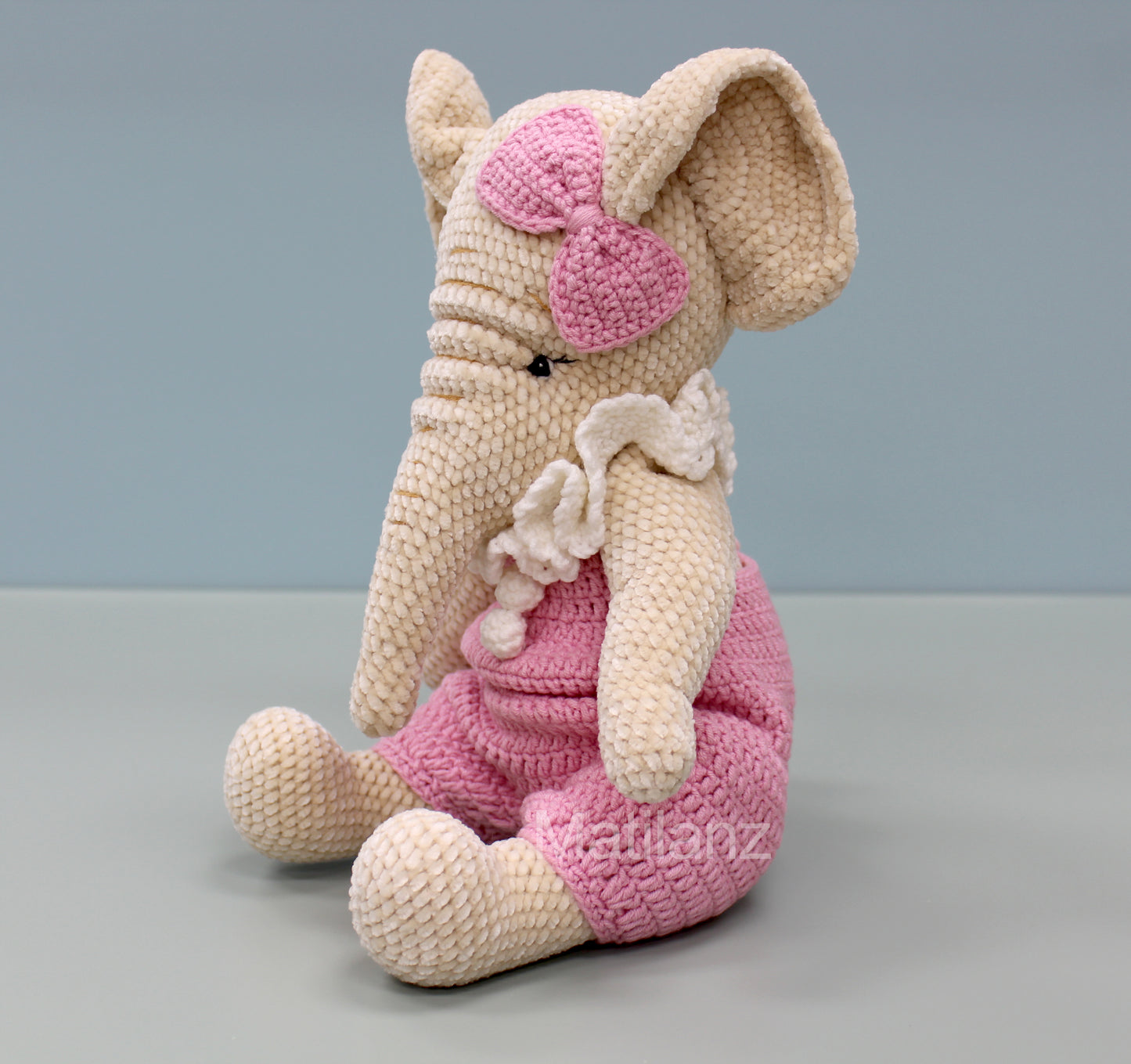 Crochet elephant in pink overalls with a bow, sitting and holding a white flower.