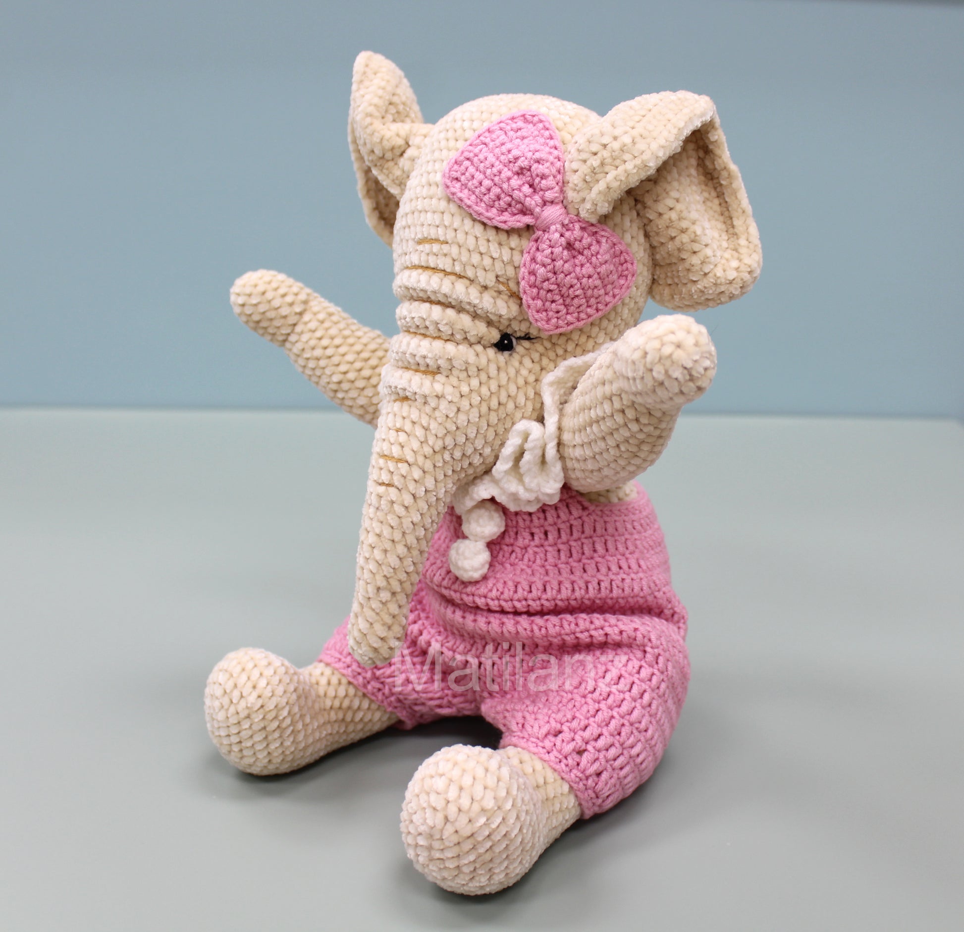 Crochet elephant in pink overalls with a bow, sitting and waving, 13 inches tall.