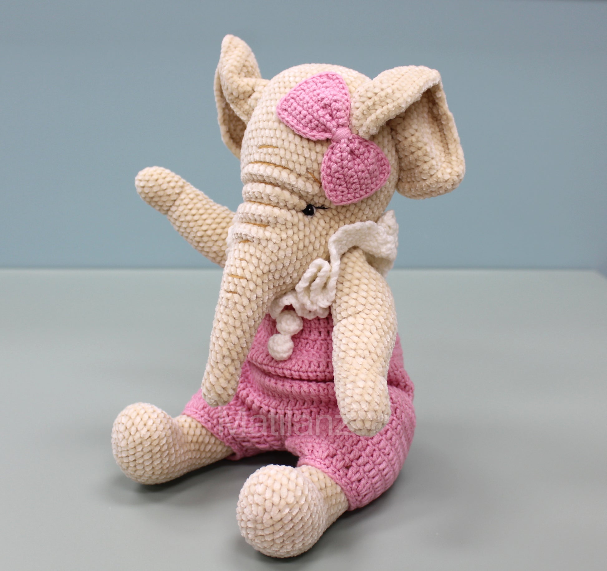 A handmade crochet elephant wearing pink overalls and a bow, sitting with one arm raised.