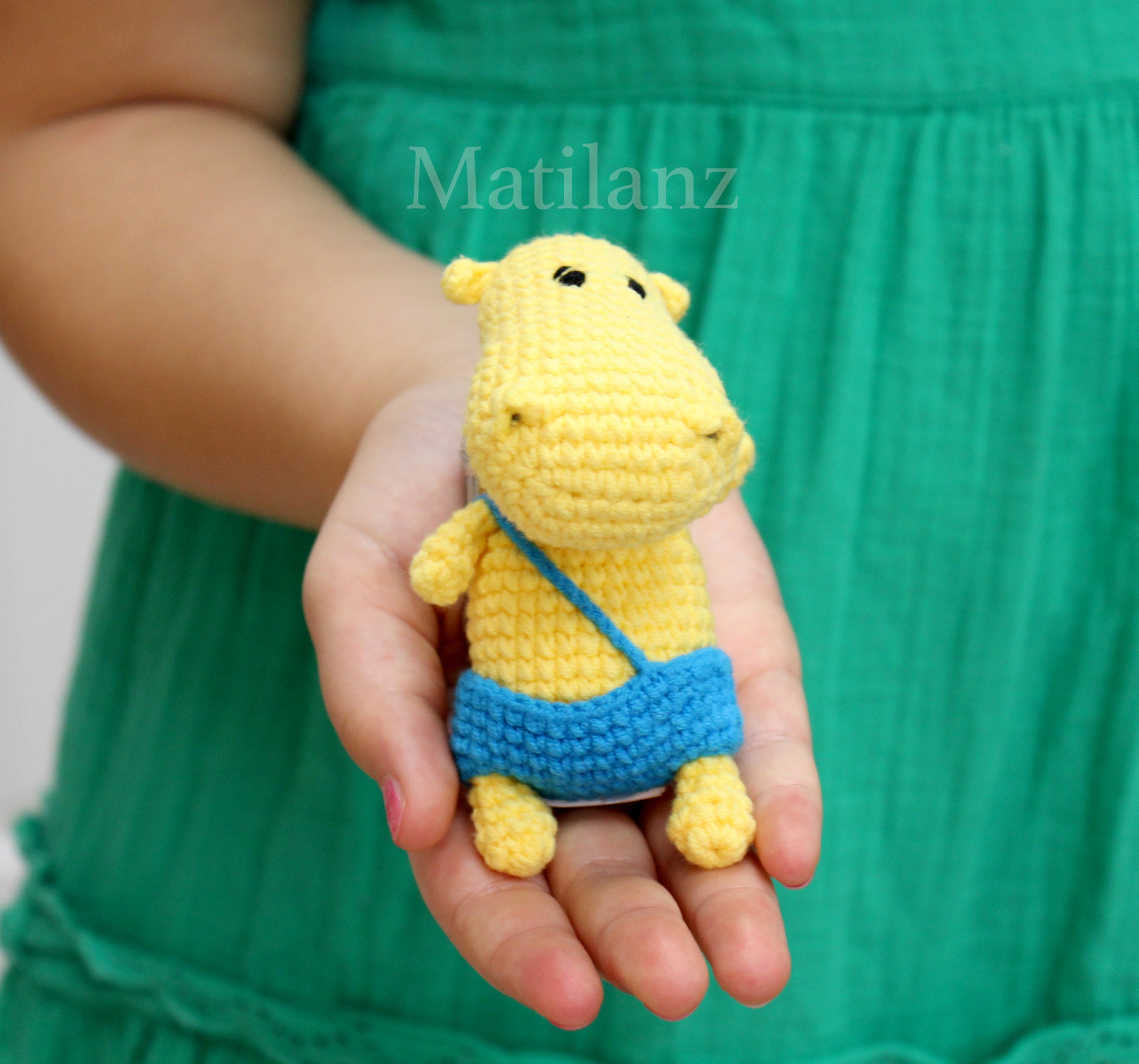 A yellow handmade crochet hippo keychain with blue overalls, held in a hand.