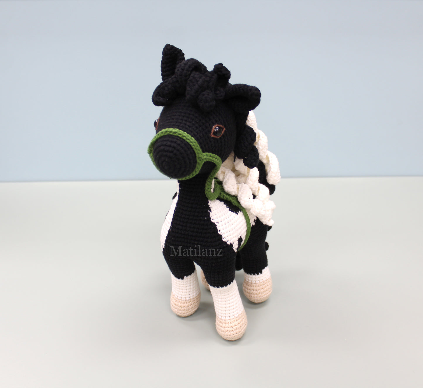 A handmade crochet paint horse plush toy with black and white stripes, green bridle, and tan hooves.