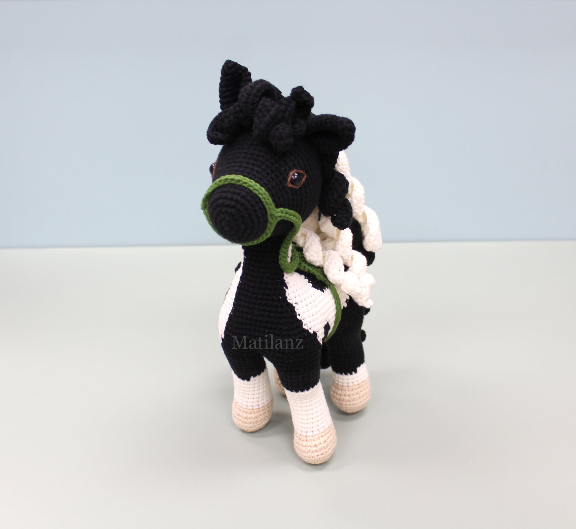 A handmade crochet paint horse plush toy with black and white stripes, green bridle, and tan hooves.