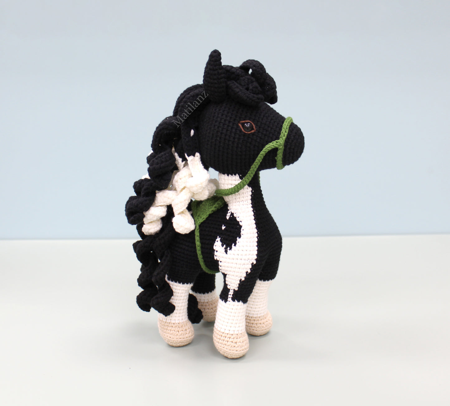 A handmade crochet paint horse plush toy with black and white patterns, green bridle, and flowing mane.