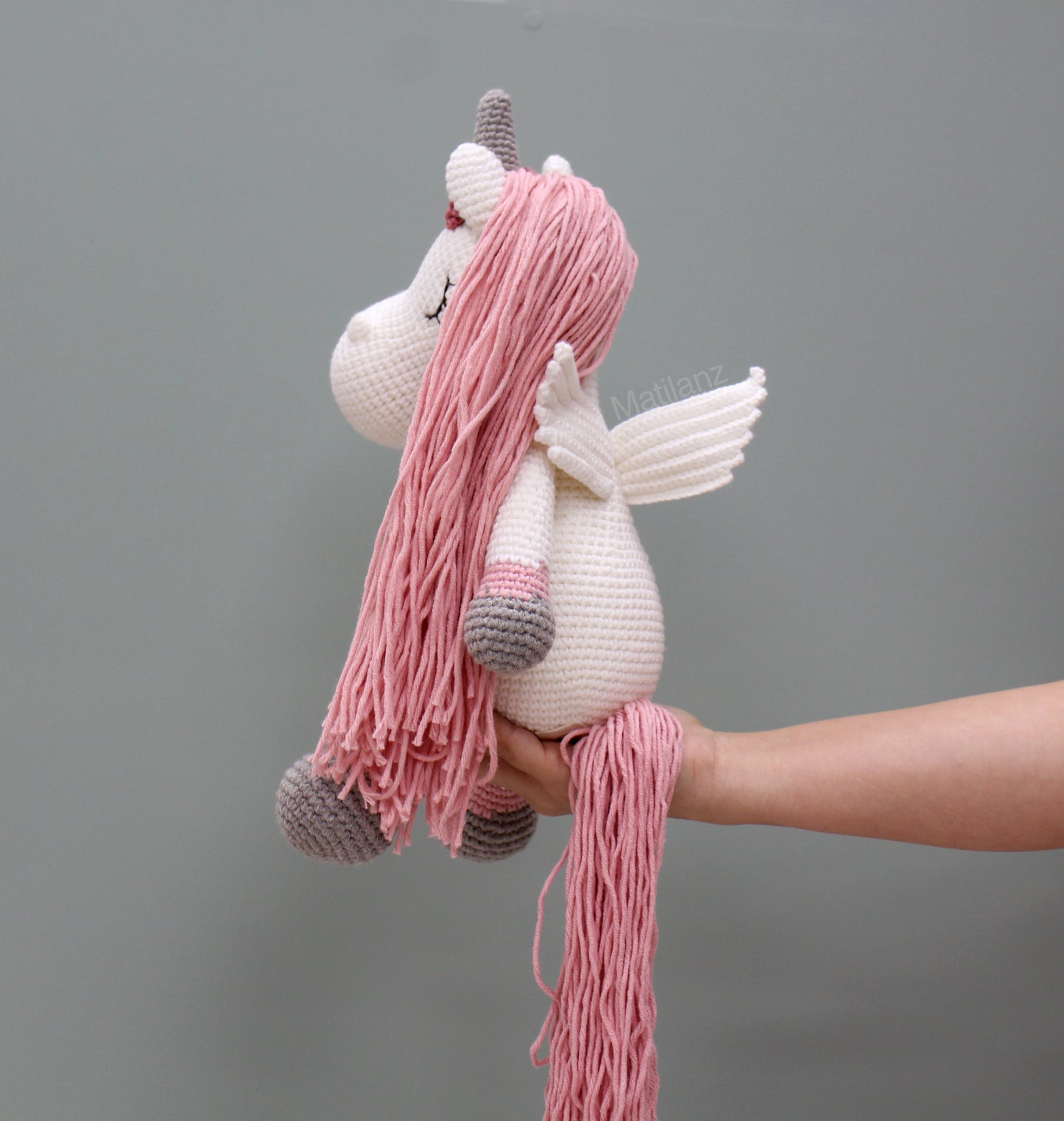 A handmade crochet unicorn with pink mane, wings, and gray hooves.