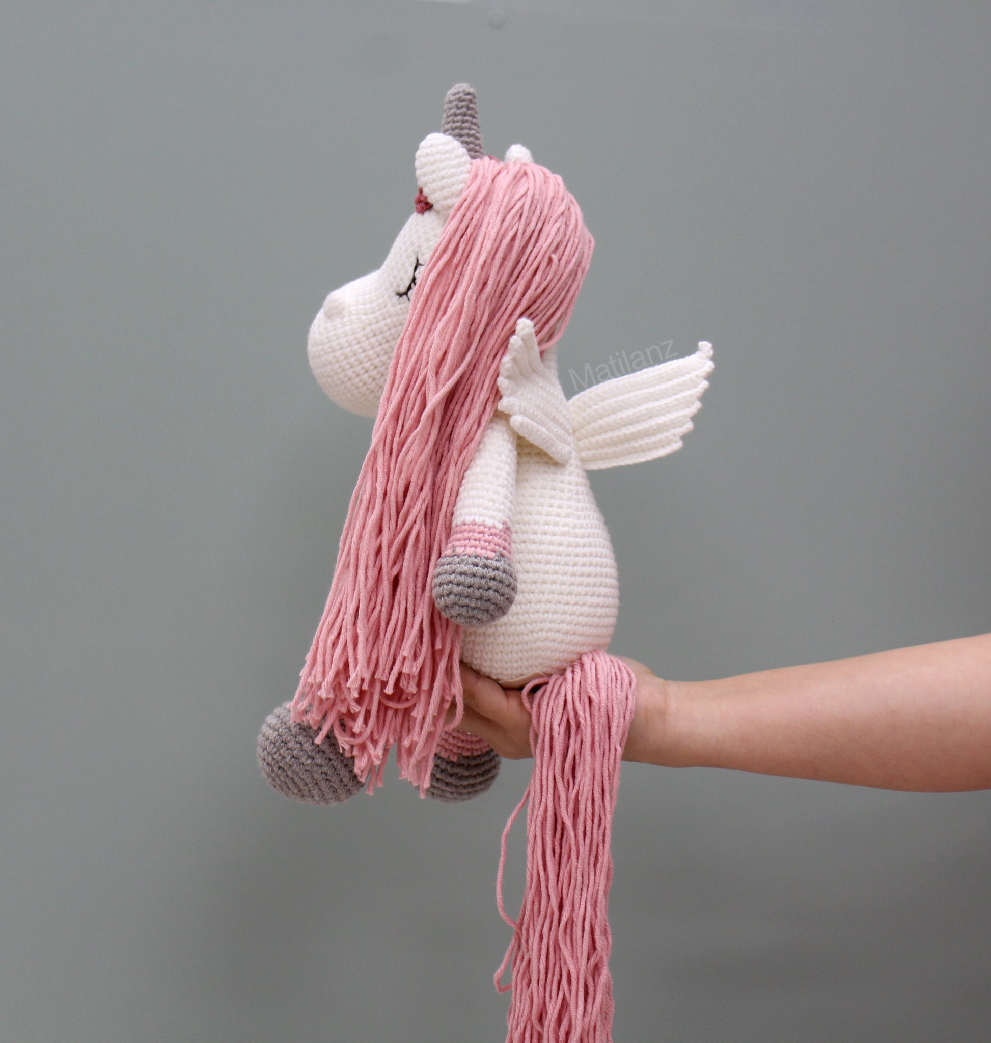 A handmade crochet unicorn with pink mane, wings, and gray hooves.
