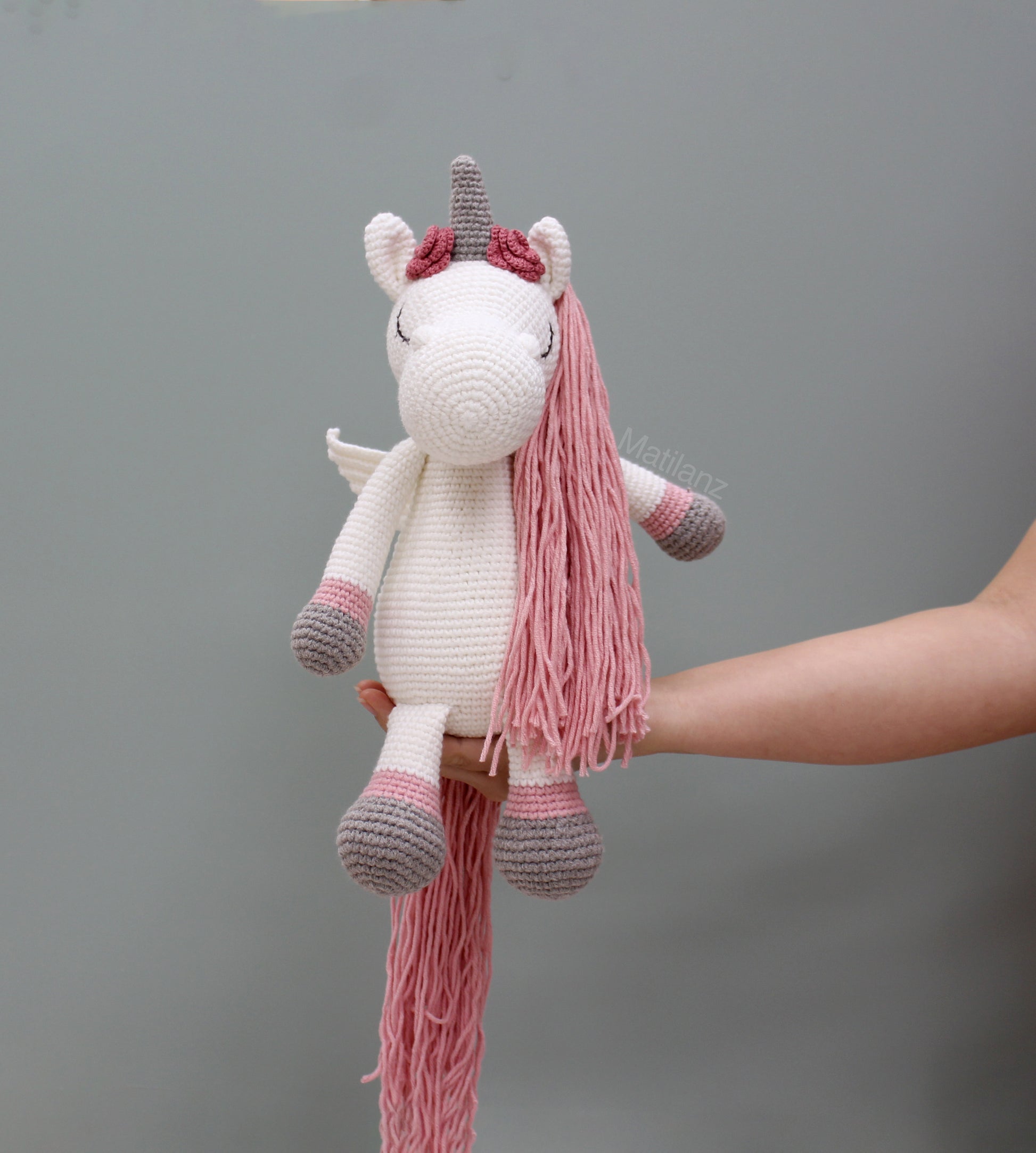 A handmade crocheted unicorn with wings, pink mane and tail, white body, and gray horn.