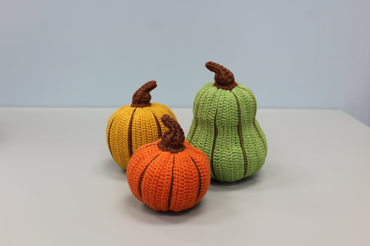 Three crocheted pumpkins in orange, yellow, and green on a light gray background