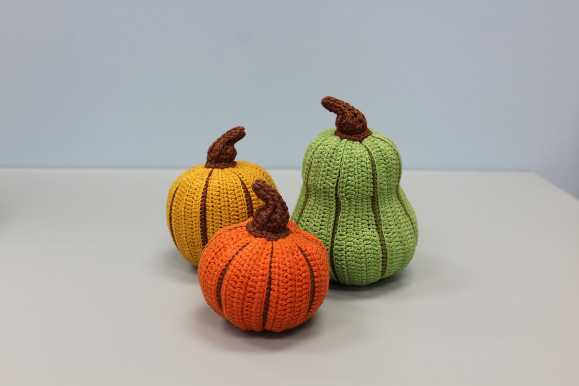 Three crocheted pumpkins in orange, yellow, and green on a light gray background