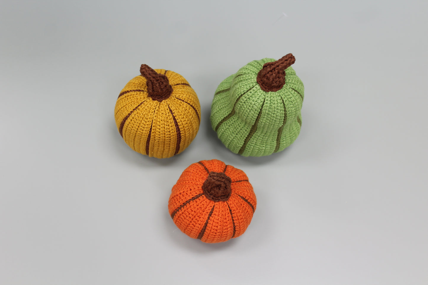 Three crocheted pumpkins in orange, yellow, and green on a light gray background