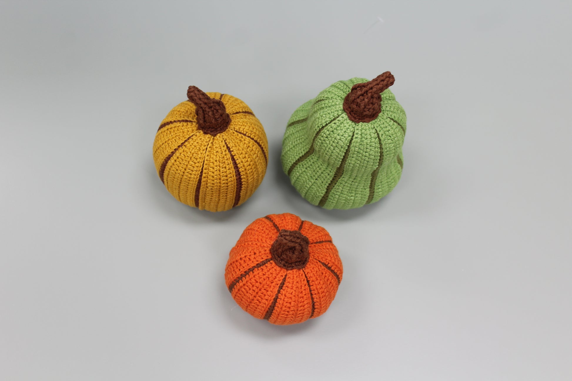 Three crocheted pumpkins in orange, yellow, and green on a light gray background
