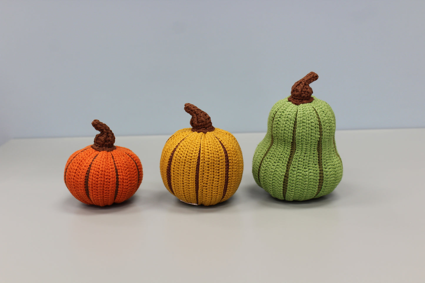 Three crocheted pumpkins in orange, yellow, and green on a light gray background