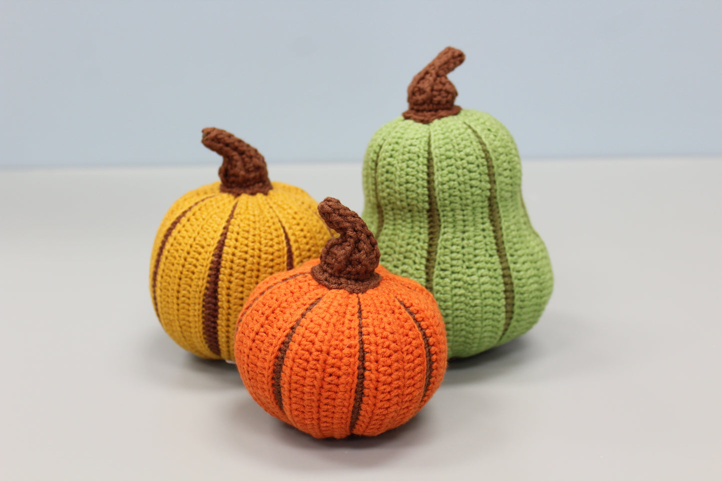 Three crocheted pumpkins in orange, yellow, and green on a light gray background