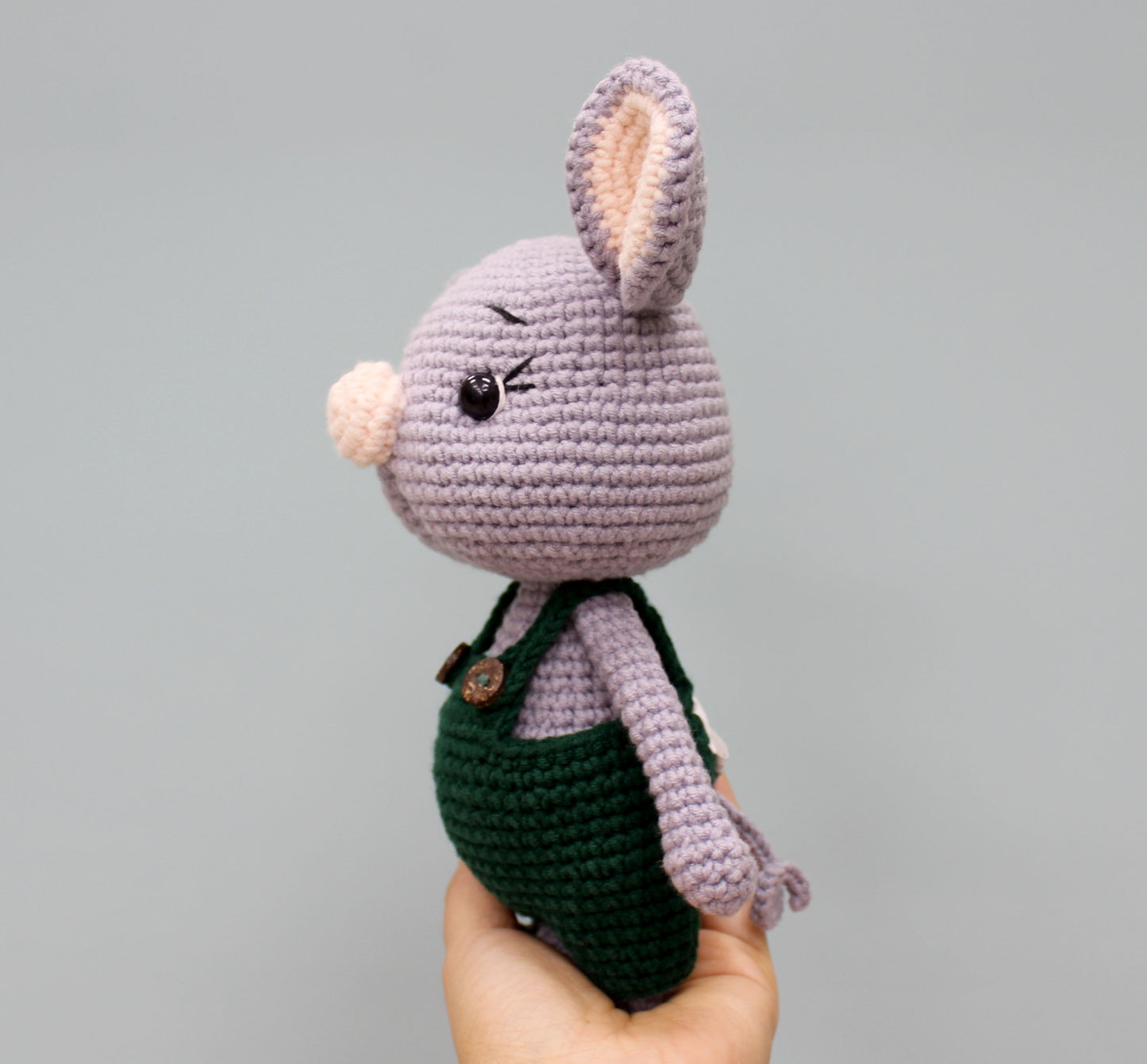 Cropped view of a handmade 7" Christmas mouse stuffed animal, featuring purple crochet material, black eyes, and a green overall outfit.