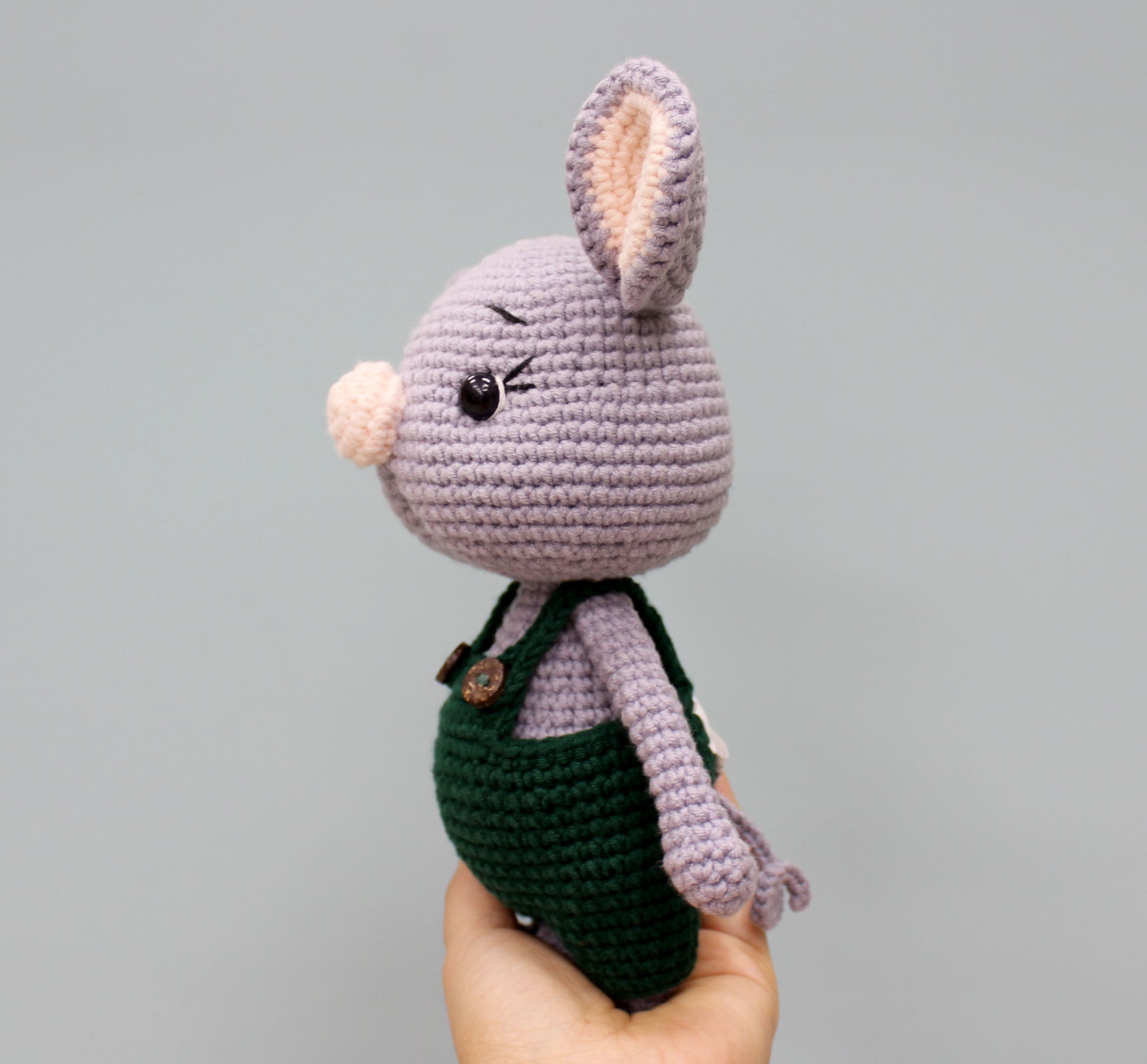 Cropped view of a handmade 7" Christmas mouse stuffed animal, featuring purple crochet material, black eyes, and a green overall outfit.