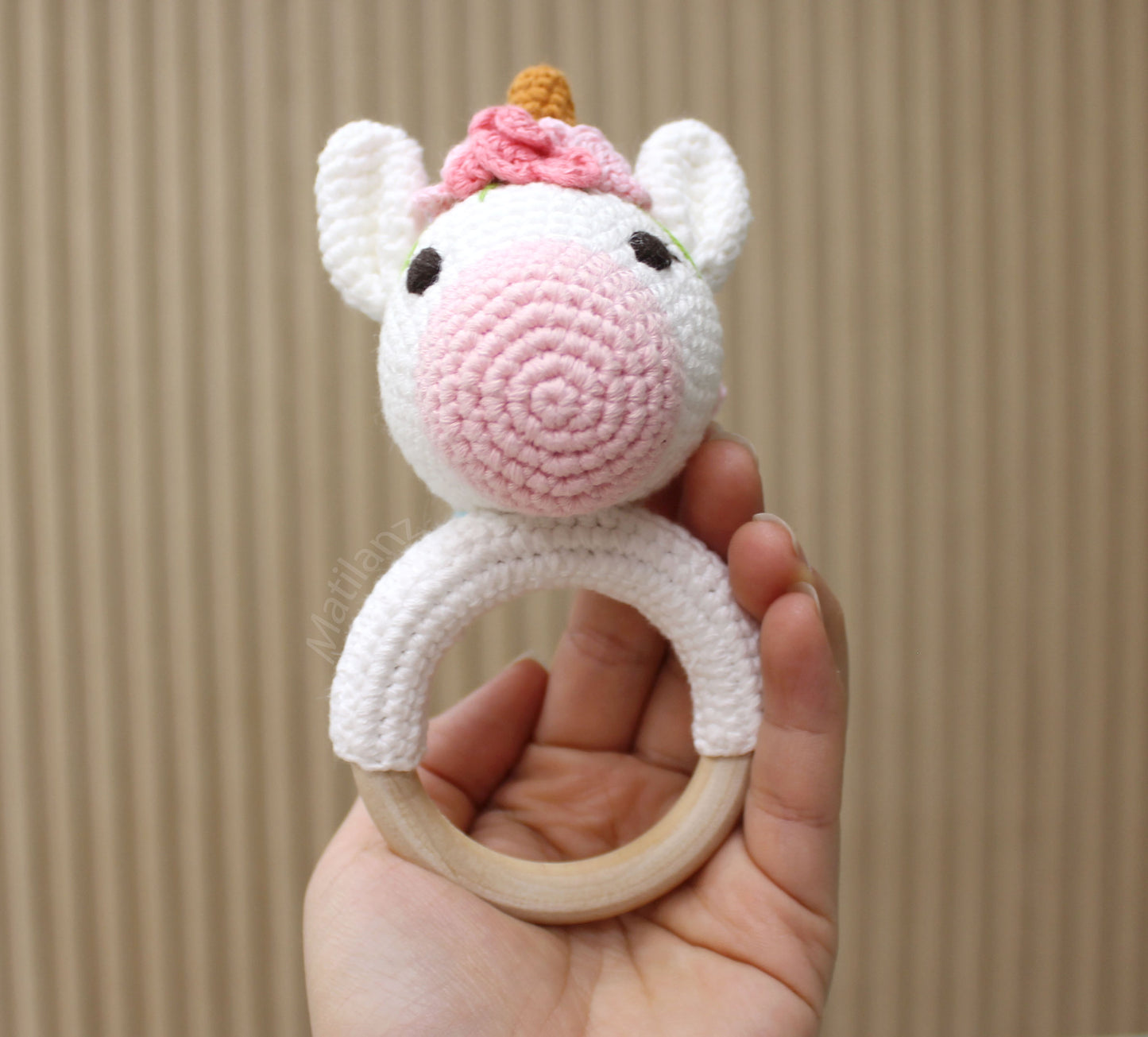 "Handmade crochet unicorn rattle toy with white body, pink mane, and wooden ring, perfect for babies."