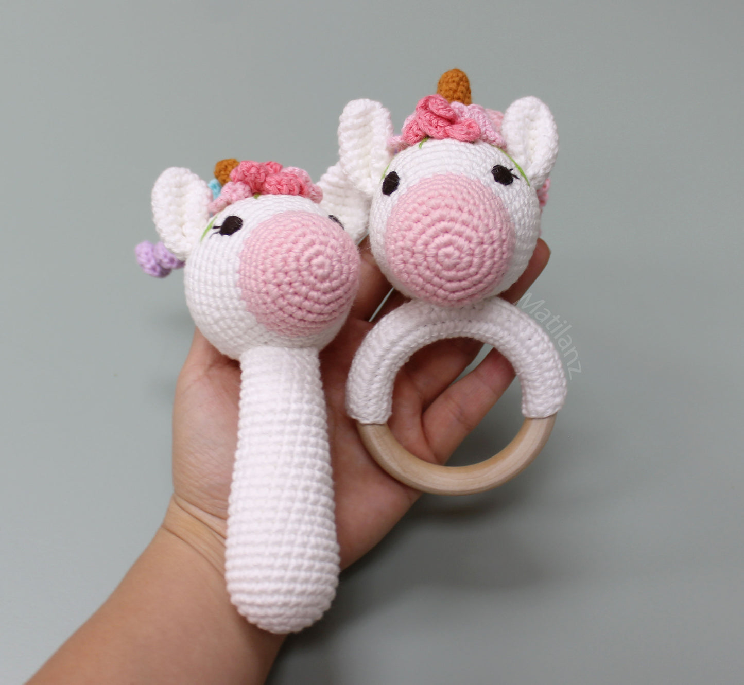 Handmade crochet unicorn baby toy with pink accents, a wooden teether ring, and a looped handle.