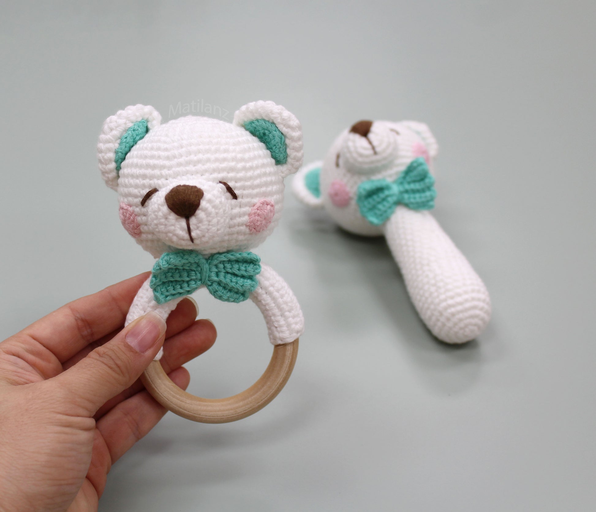 Handmade crochet bear rattle toy in white with teal accents, featuring a wooden ring and bow tie.