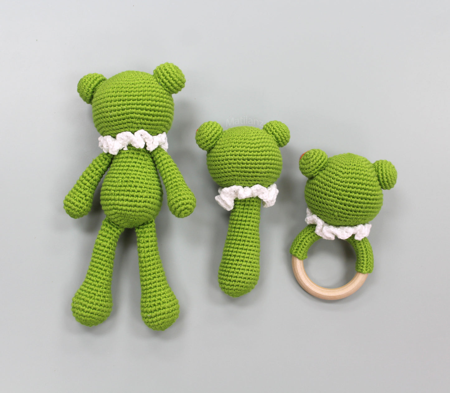 A handmade green crochet frog baby toy set, featuring a three-piece sensory collection with white ruffled accents and a wooden ring.