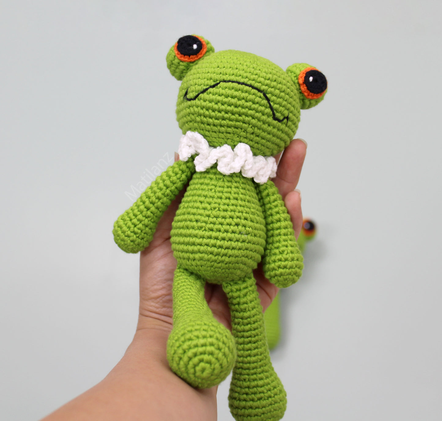A handmade green frog plush with a white collar, orange-rimmed eyes, and a soft, textured crochet finish.