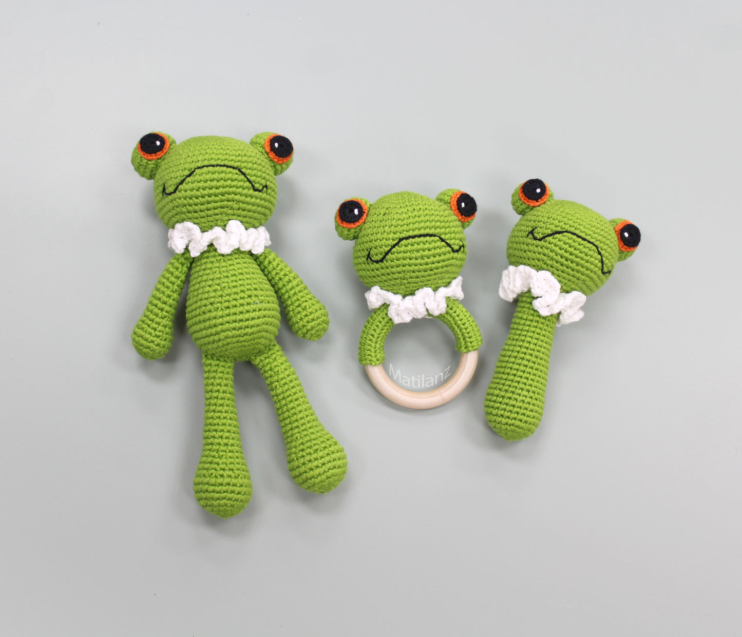 A handmade green crochet frog baby toy set with white collar, large eyes, and a wooden ring teether.